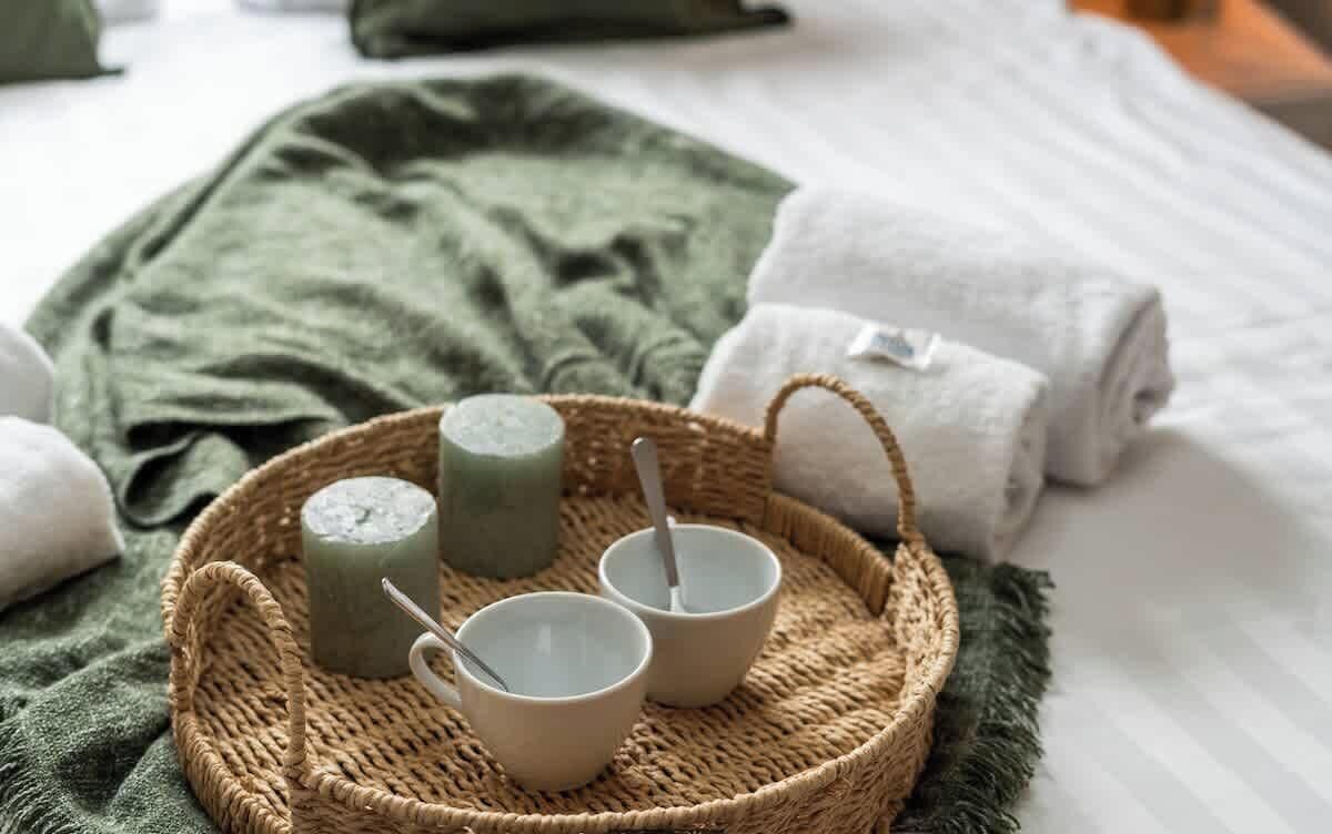 Een bed met een groene deken, twee opgerolde handdoeken en een geweven dienblad met twee bekers en een stuk zeep.