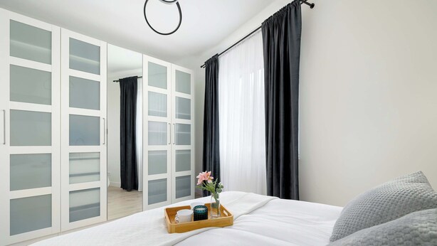 Une chambre à coucher avec un lit, un plateau en bois sur lequel se trouvent des tasses et un vase, une armoire avec des portes en verre dépoli et des rideaux noirs.