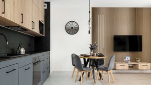 Une cuisine moderne dotée d’un four encastré, d’un coin repas avec une table ronde et quatre chaises, d’une horloge murale et d’un téléviseur à écran plat fixé sur un mur recouvert de panneaux en bois.
