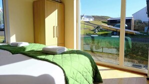 Une chambre à coucher avec un lit, une couette verte, une armoire en bois et une grande fenêtre donnant sur une maison et un bateau.