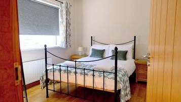Um quarto com estrutura de cama de metal, uma mesa de cabeceira, uma janela com cortina e um radiador.