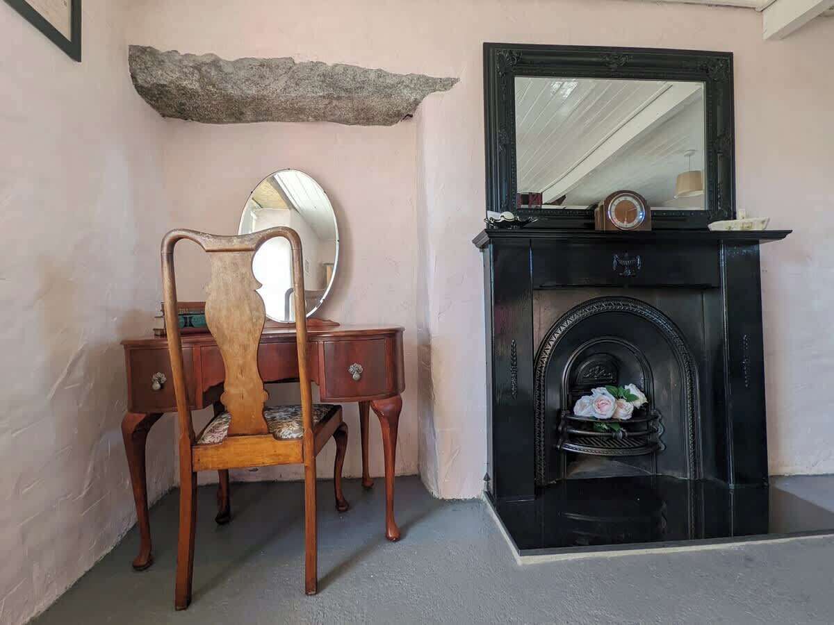 A room with a wooden chair, a desk, a mirror, a fireplace with a flower arrangement, and a stone shelf above the fireplace.
