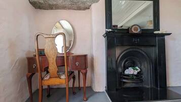 A room with a wooden chair, a desk, a mirror, a fireplace with a flower arrangement, and a stone shelf above the fireplace.