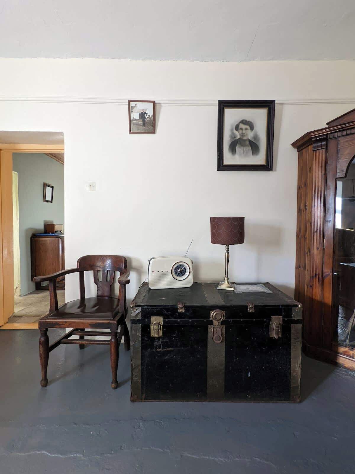 Um quarto com uma cadeira de madeira, um rádio antigo, uma luminária e um baú.