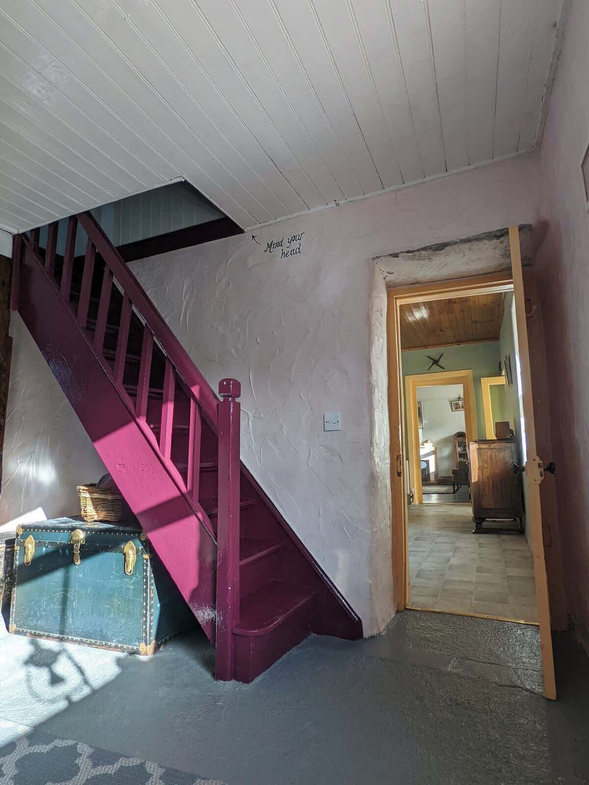 A staircase with a red handrail leading to a room with a wooden door and a wall-mounted light switch.
