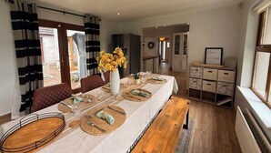A dining area with a long table set for six, a vase of flowers, and a view of the kitchen through an open door. - Modern 4 Bed,3 Bath Detached Home with Sea View (Quigley's Point)