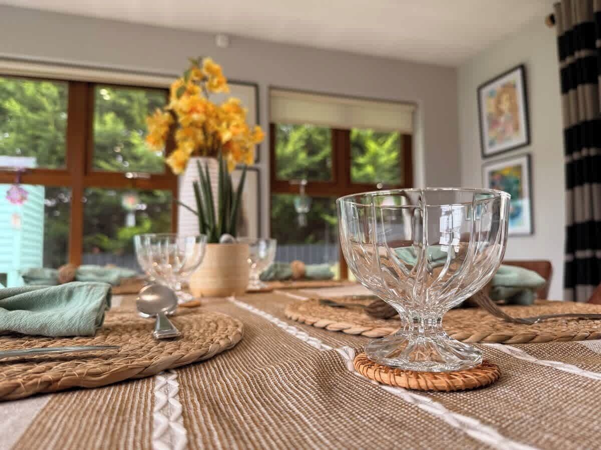 A dining table with a clear glass bowl, a spoon, and a vase with yellow flowers.