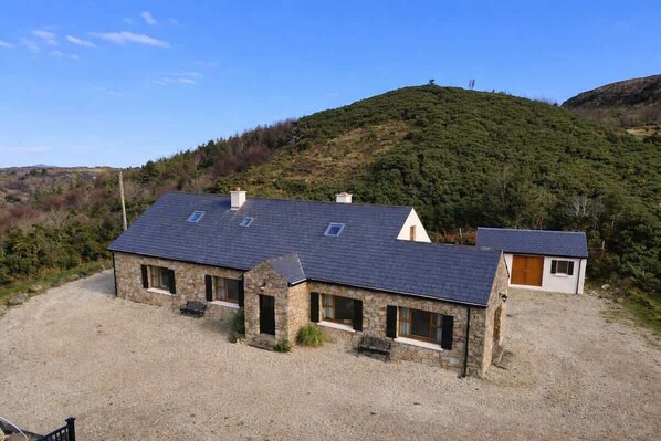 A stone building with a sloped roof, a gravel driveway, and a hillside in the background.