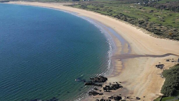 Vista aérea de uma área costeira com praia arenosa, afloramentos rochosos e campos verdes.