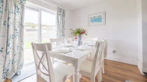 Un coin repas avec une table blanche dressée pour quatre personnes, des chaises blanches, un vase rempli de fleurs et une vue sur l’extérieur grâce à une porte vitrée.