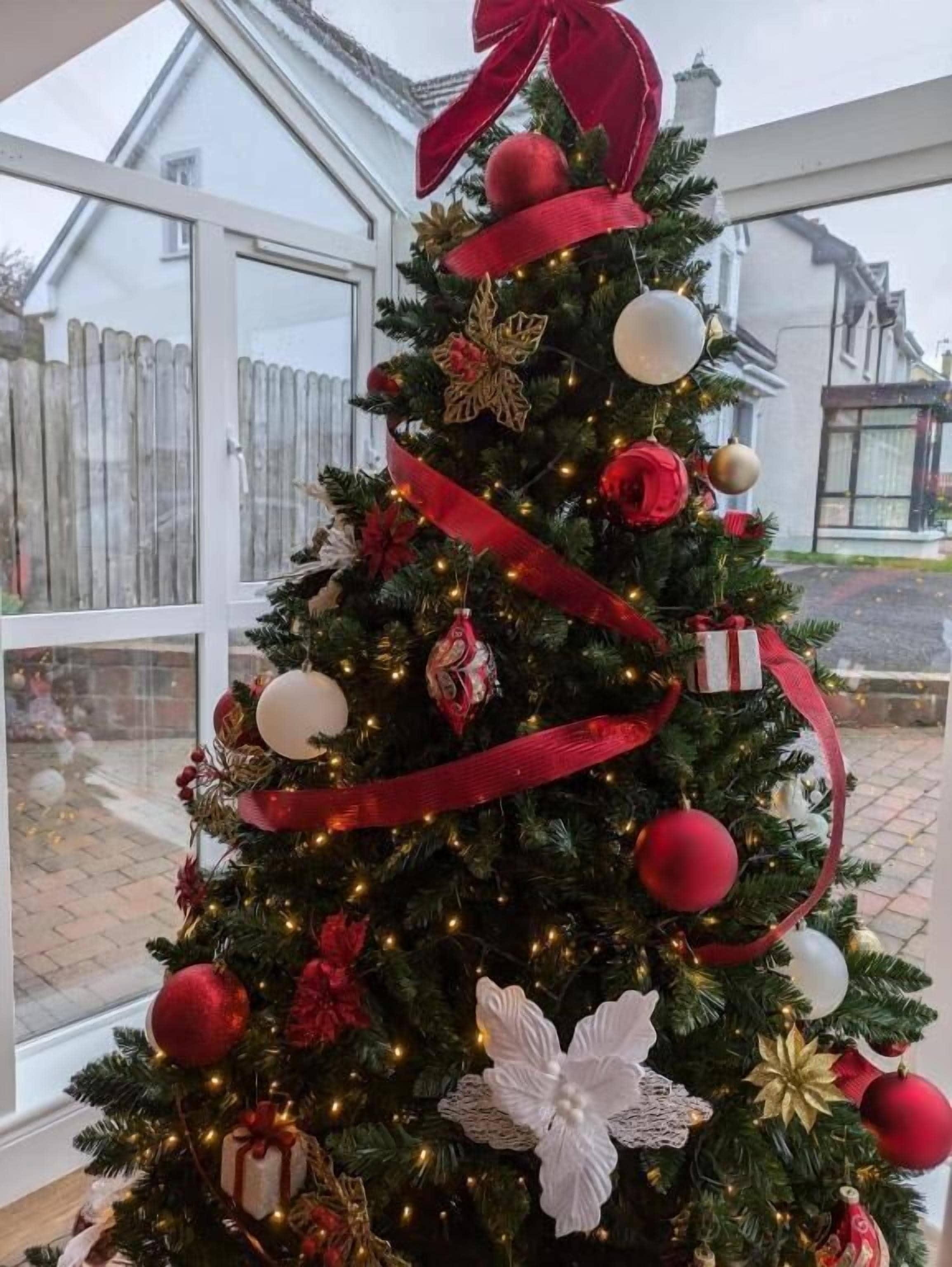 A Christmas tree decorated with ornaments and lights, placed indoors with a view of a house outside.