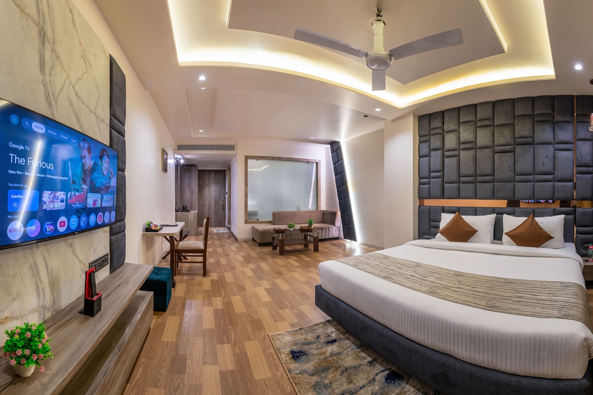 Habitación de hotel moderna con una cama grande, televisor montado en la pared, ventilador de techo y una zona de estar con una mesa y sillas.