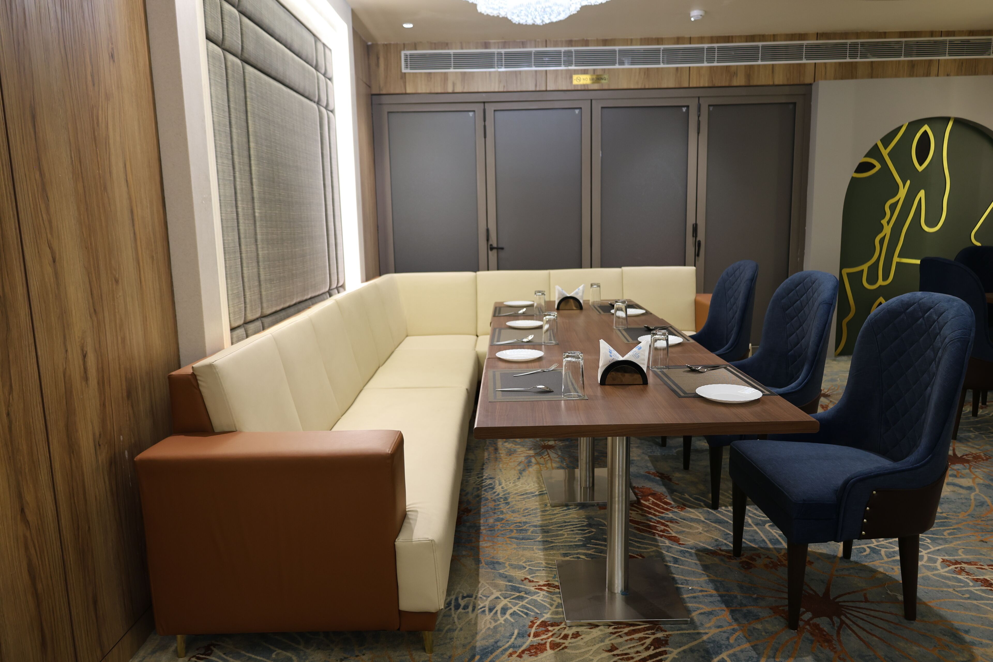 A hotel dining area with a long table, blue upholstered chairs, a cream-colored sofa, and a patterned carpet.