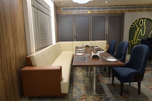 Un comedor de hotel con una mesa larga, sillas tapizadas en azul, un sofá color crema y una alfombra estampada.