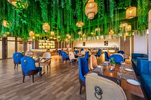 A restaurant with hanging plants and wicker lamps, wooden flooring, and blue upholstered chairs.