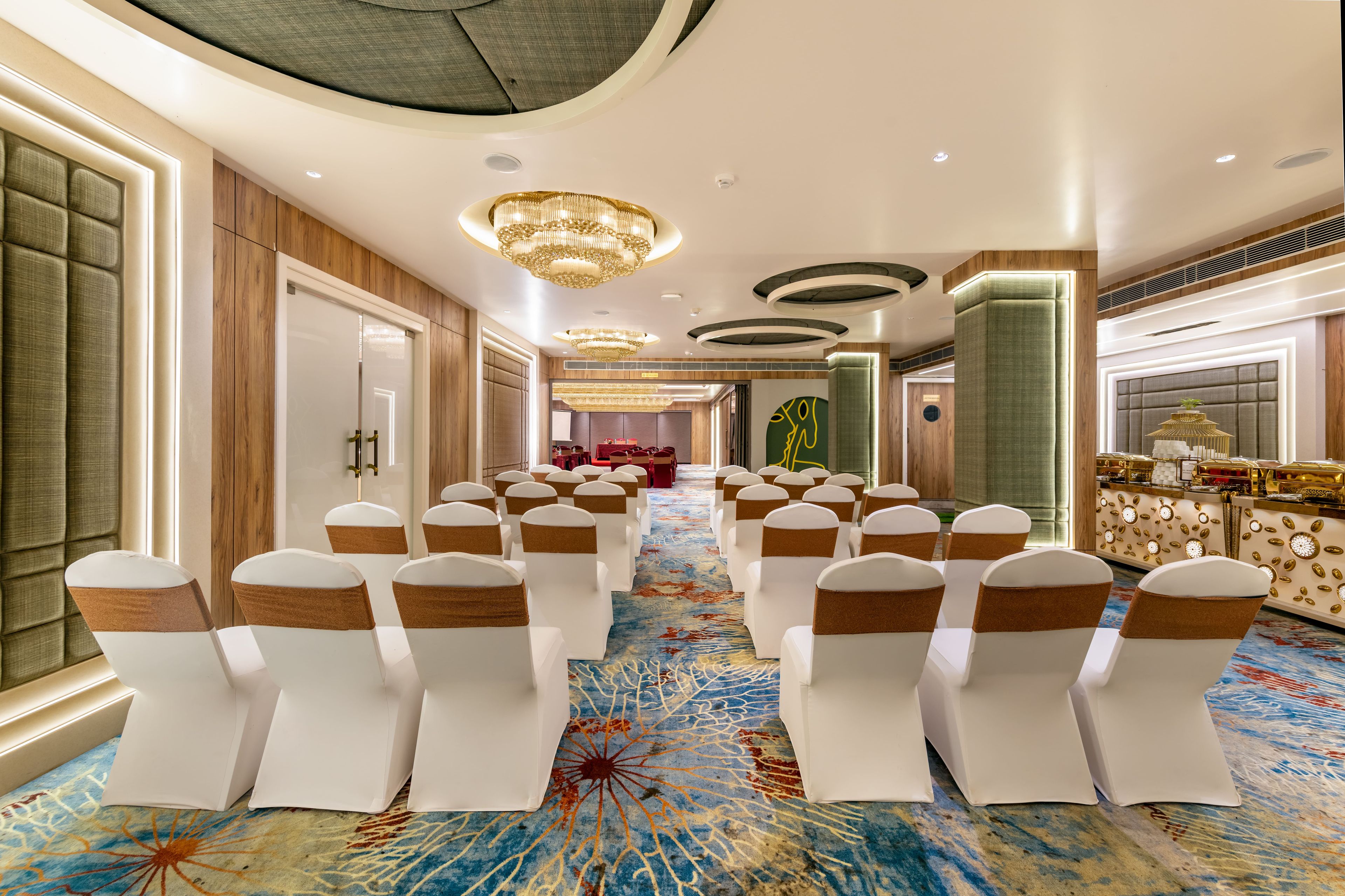 Uma sala de conferências de hotel com fileiras de cadeiras brancas, carpete estampado e um candelabro.