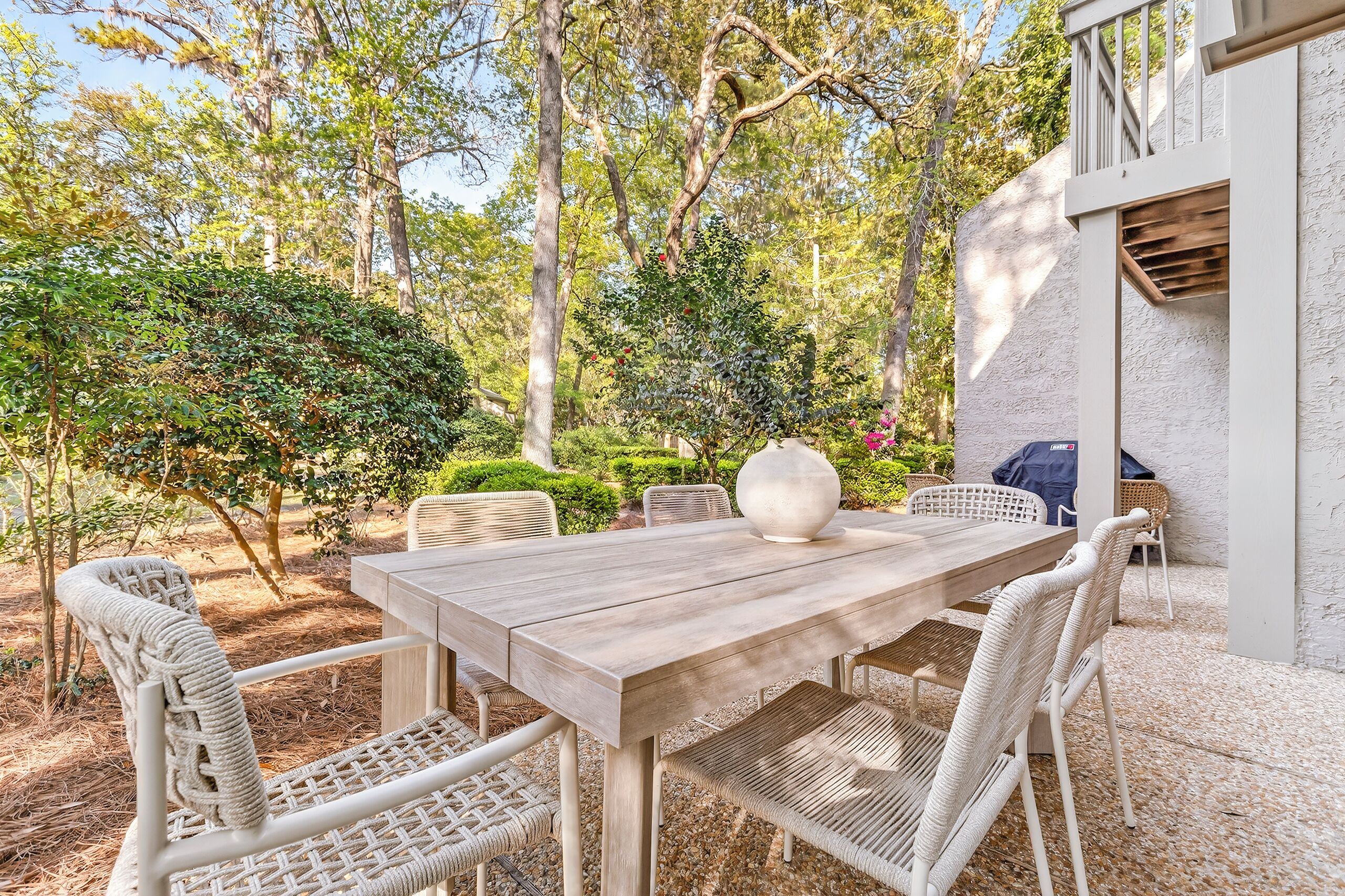 An outdoor patio with a wooden table and wicker chairs, surrounded by greenery and trees.