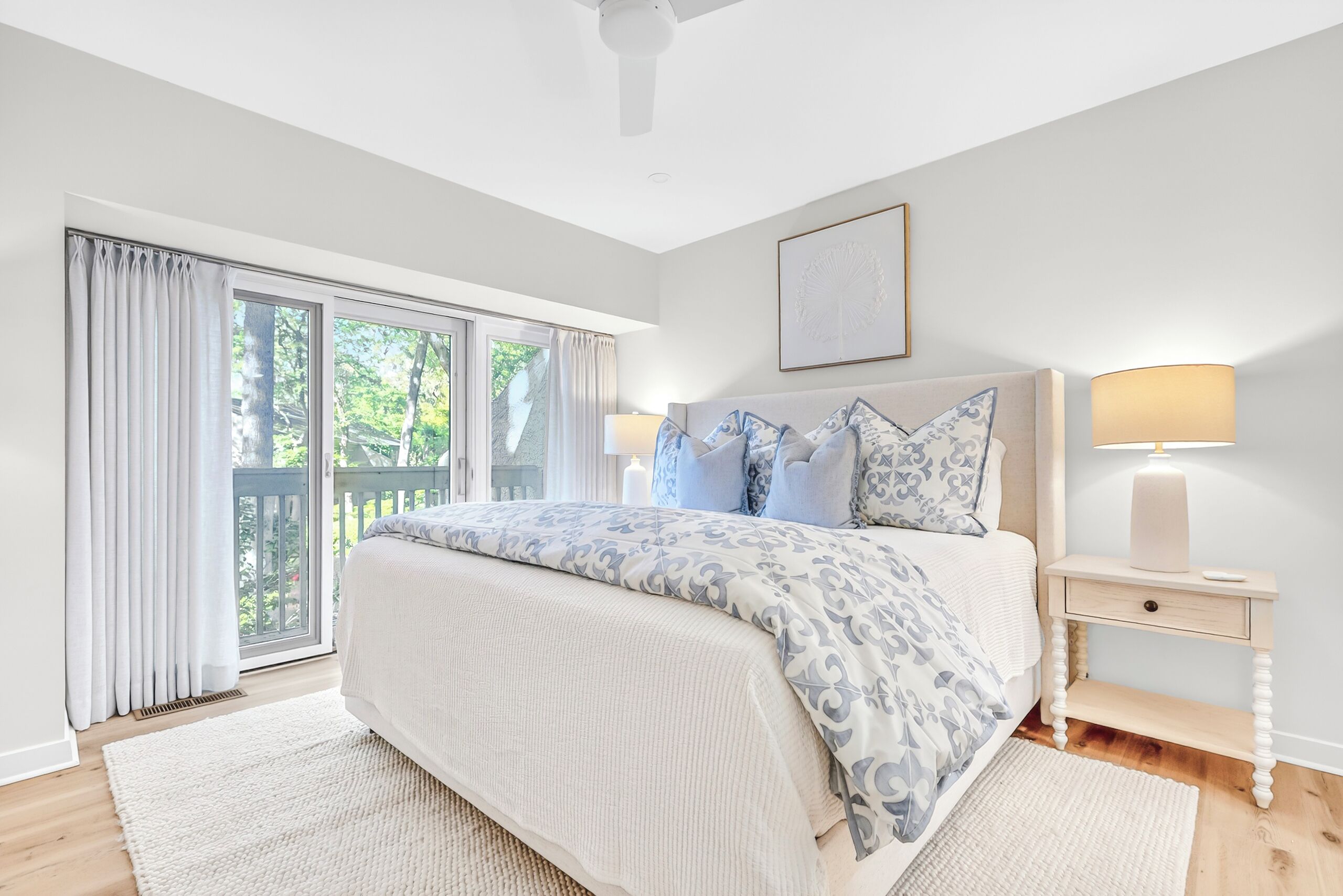 Une chambre avec un grand lit, une table de chevet avec une lampe, un ventilateur de plafond et une vue sur l’extérieur grâce à une porte-fenêtre coulissante.