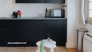 Une cuisine moderne avec des meubles noirs, un four à micro-ondes et un évier. On y trouve deux tables rondes blanches, dont l’une est ornée d’un objet décoratif vert. Un canapé gris se trouve au premier plan.