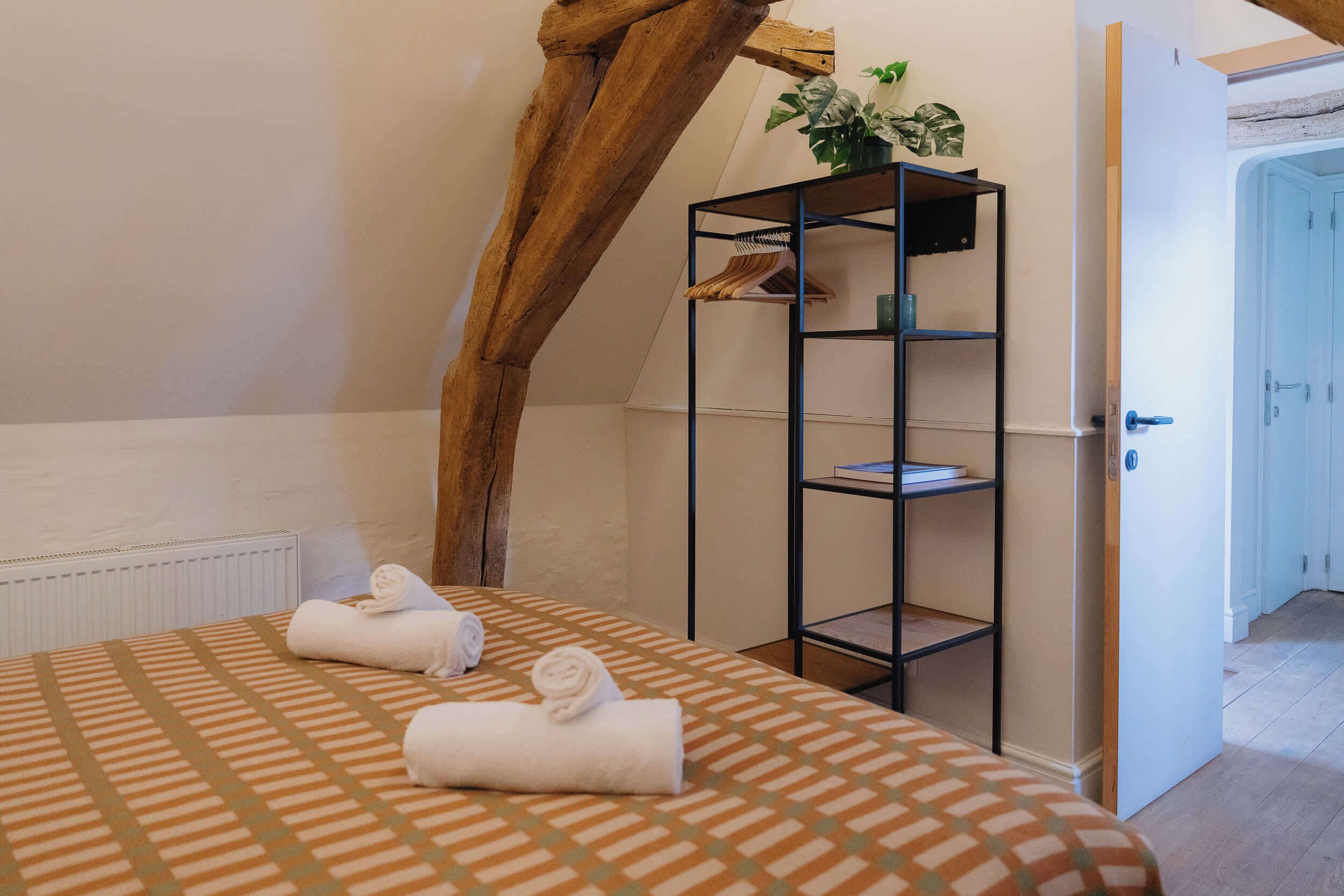 Een slaapkamer met een bed, handdoeken, een metalen zwarte plank, een potplant en een deur naar een andere kamer.