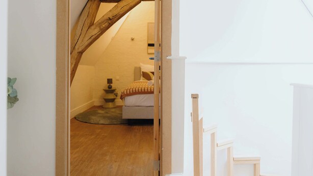 Une vue intérieure d’une pièce avec un plafond en bois, un lit recouvert d’une couvre-lit à motifs, une petite table avec une lampe et un panier pour animal de compagnie.
