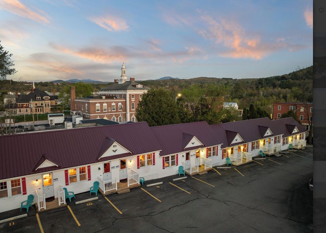 A motel with a row of rooms, a parking lot, and a large building in the background.