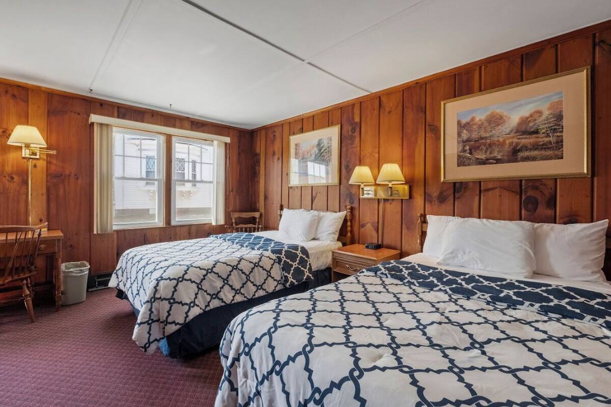 A room with two beds, wooden walls, a desk, and two lamps.