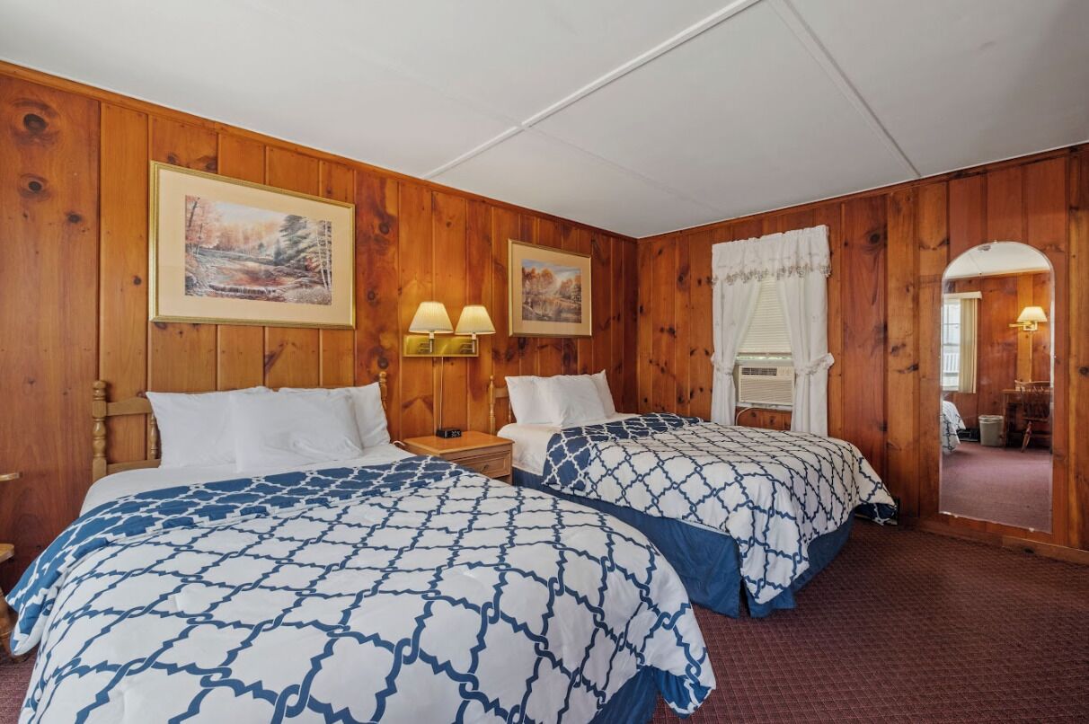 A room with wooden paneling, two beds with patterned bedding, wall-mounted lamps, and framed pictures.