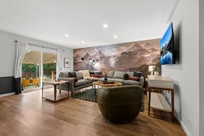 Un salon moderne avec un canapé, une table basse, un téléviseur et une fresque représentant des montagnes.