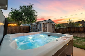 A hot tub in a backyard with a wooden fence, a small shed, and a house in the background.