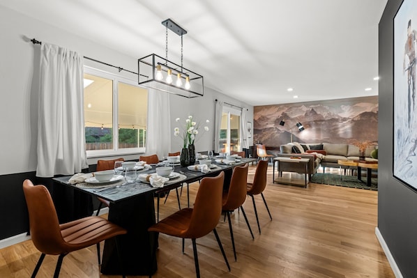 Un coin repas avec une longue table dressée pour quatre personnes, entourée de chaises, et un lustre moderne au-dessus.