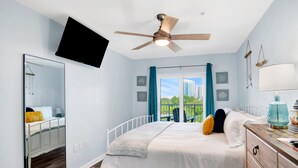 A bedroom with a bed, a ceiling fan, a TV mounted on the wall, and a view of the outdoors. - Beach Access 2BR @Grand Caribbean • Sleeps 6 • Views, Balcony & Pool (Orange Beach)