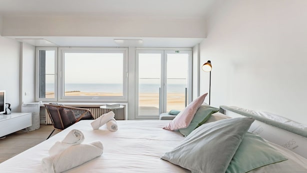 Een hotelkamer met een bed, een stoel, een lamp en uitzicht op het strand.