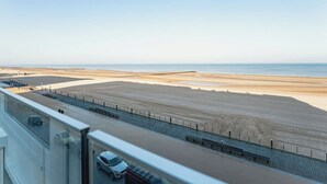 Uitzicht op de kust vanaf een hoog punt, met een zandstrand, een verharde promenade en een verre waterpartij.