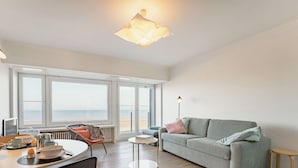 Een moderne woonkamer met een bank, eettafel, stoelen en grote ramen met uitzicht op de zee.