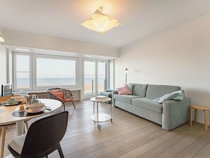 Ein modernes Wohnzimmer mit einer Couch, einem Esstisch, Stühlen und großen Fenstern mit Blick aufs Meer.