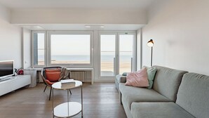 Een moderne woonkamer met een bank, een klein tafeltje, een stoel en een televisie. Grote ramen bieden uitzicht op het strand.