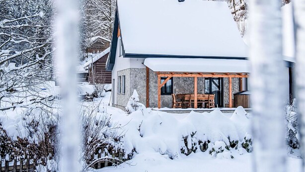 Eine schneebedeckte Lodge mit Steinaußenwand, einer Holzdiele und Blick auf einen Wald.
