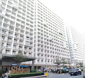 A high-rise building with multiple balconies, a sign for SMDC Shore, and a street with pedestrians and vehicles.