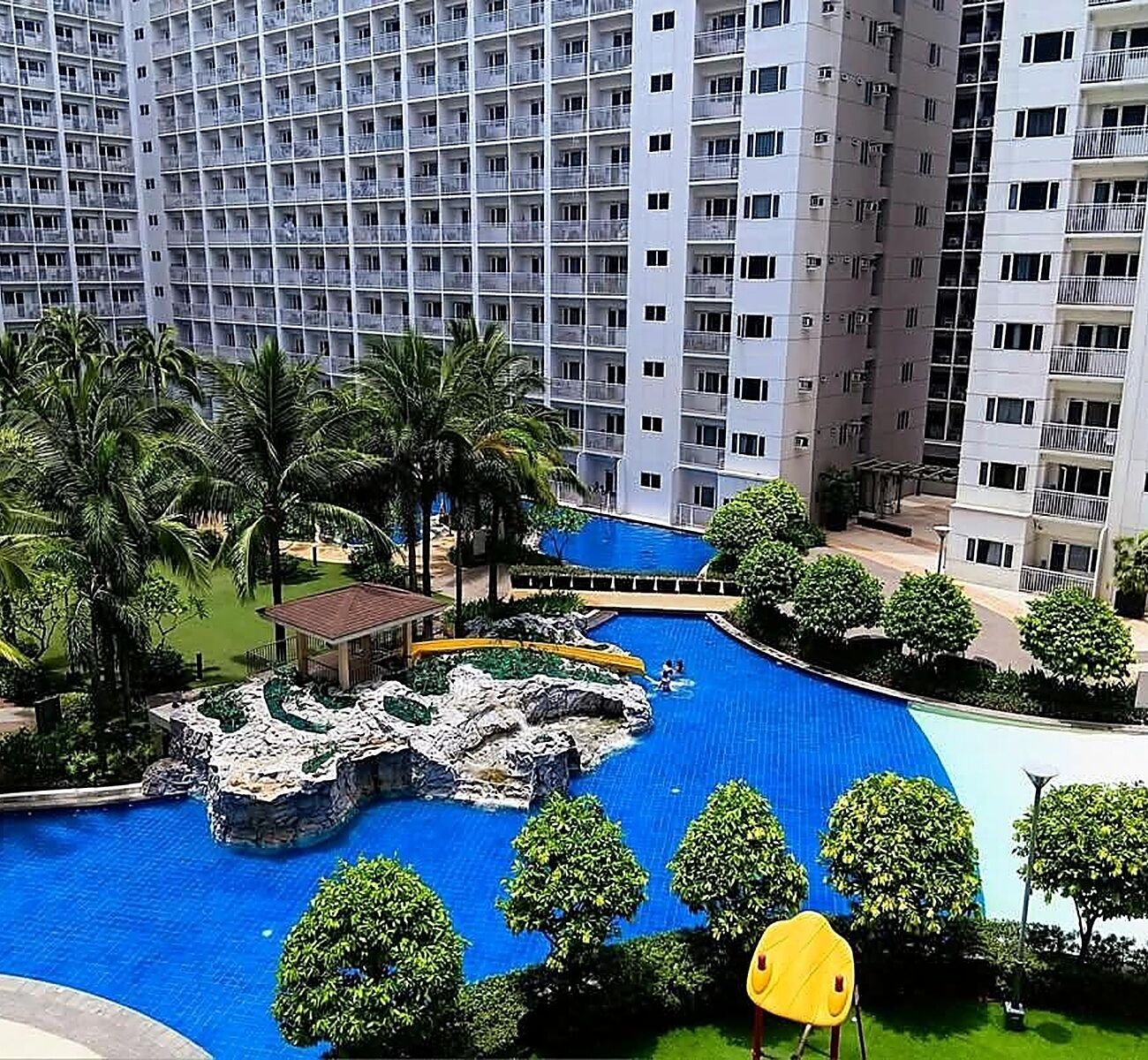 A high-rise hotel with a swimming pool, palm trees, and a rock feature.