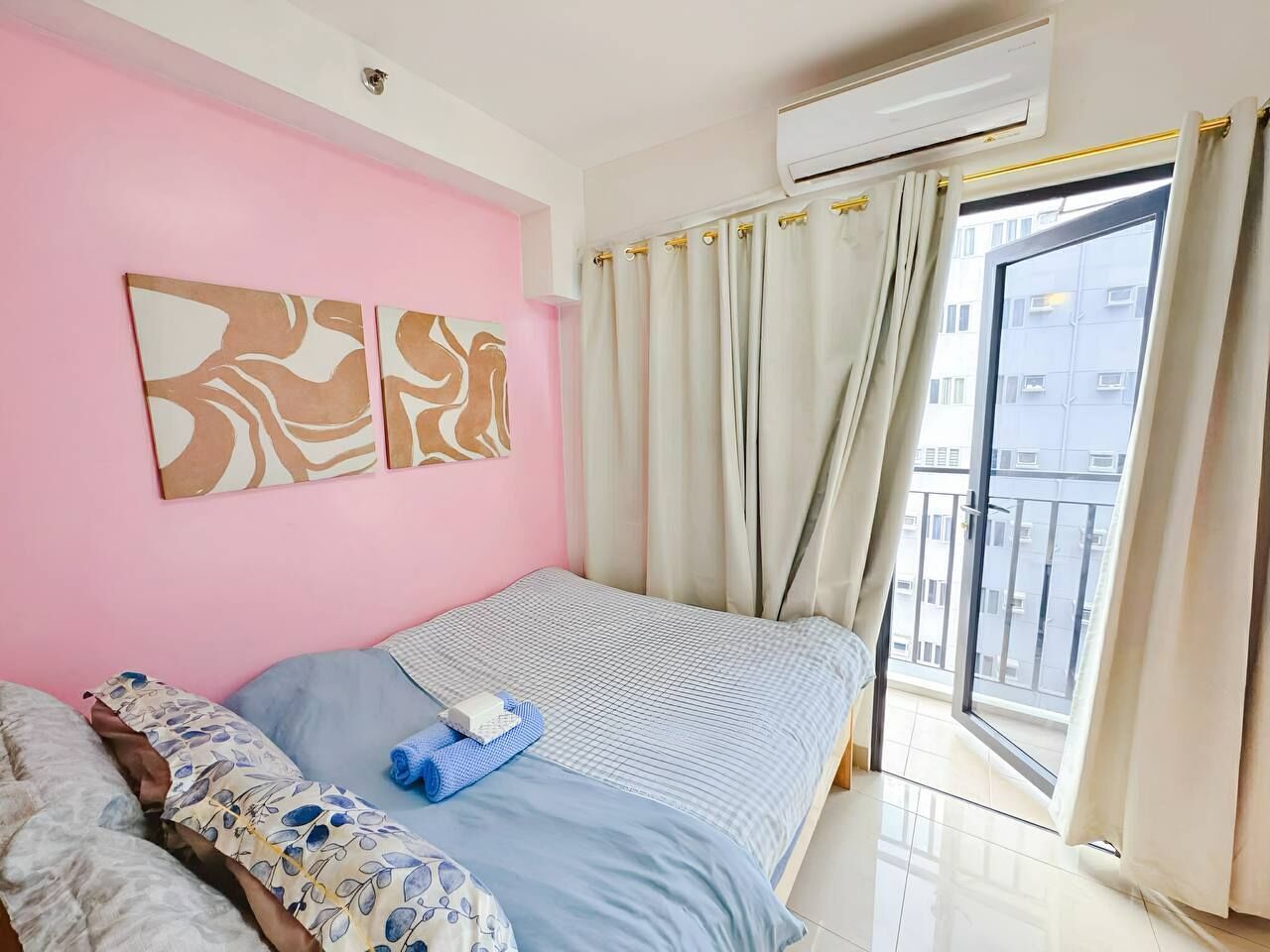 Un dormitorio con una pared rosa, una cama con colcha azul y blanca a cuadros, dos almohadas, una toalla azul y una ventana con cortinas.