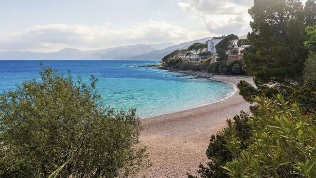 Une plage côtière aux eaux d’un bleu limpide, avec un rivage sablonneux et un bâtiment doté d’un toit en pente, situé sur une colline.