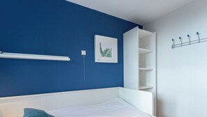 Un dormitorio con cama blanca, paredes azules, cabecera blanca, estante blanco y perchero de pared.