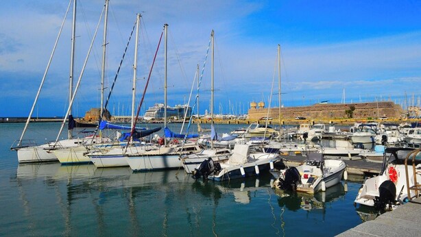Un puerto deportivo con varias embarcaciones amarradas, cielo despejado y un edificio a lo lejos.