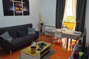 Een woonkamer met een donkere bank, een salontafel met een fles en glazen, een eethoek met een tafel gedekt voor twee personen, en een tv op een dressoir.
