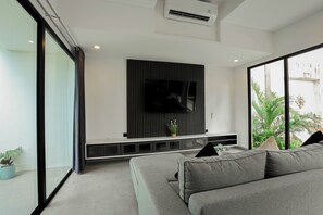 A modern living room with a grey sofa, a flat-screen TV mounted on a black panel, and a large window with a view of greenery. - D'Kenyeri Villa by Aayan (Tabanan)