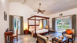 A spacious bedroom with a four-poster bed, a sitting area with a sofa and armchairs, a wooden desk, and a large window offering a view of the outdoors. - ELIVAAS The Aravalli Reserve Isley Villa (Faridabad)