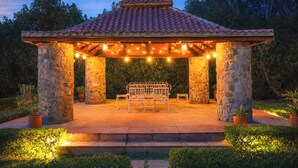 A stone gazebo with a tiled roof, surrounded by potted plants, and featuring a bench and table setup. - ELIVAAS The Aravalli Reserve Isley Villa (Faridabad)