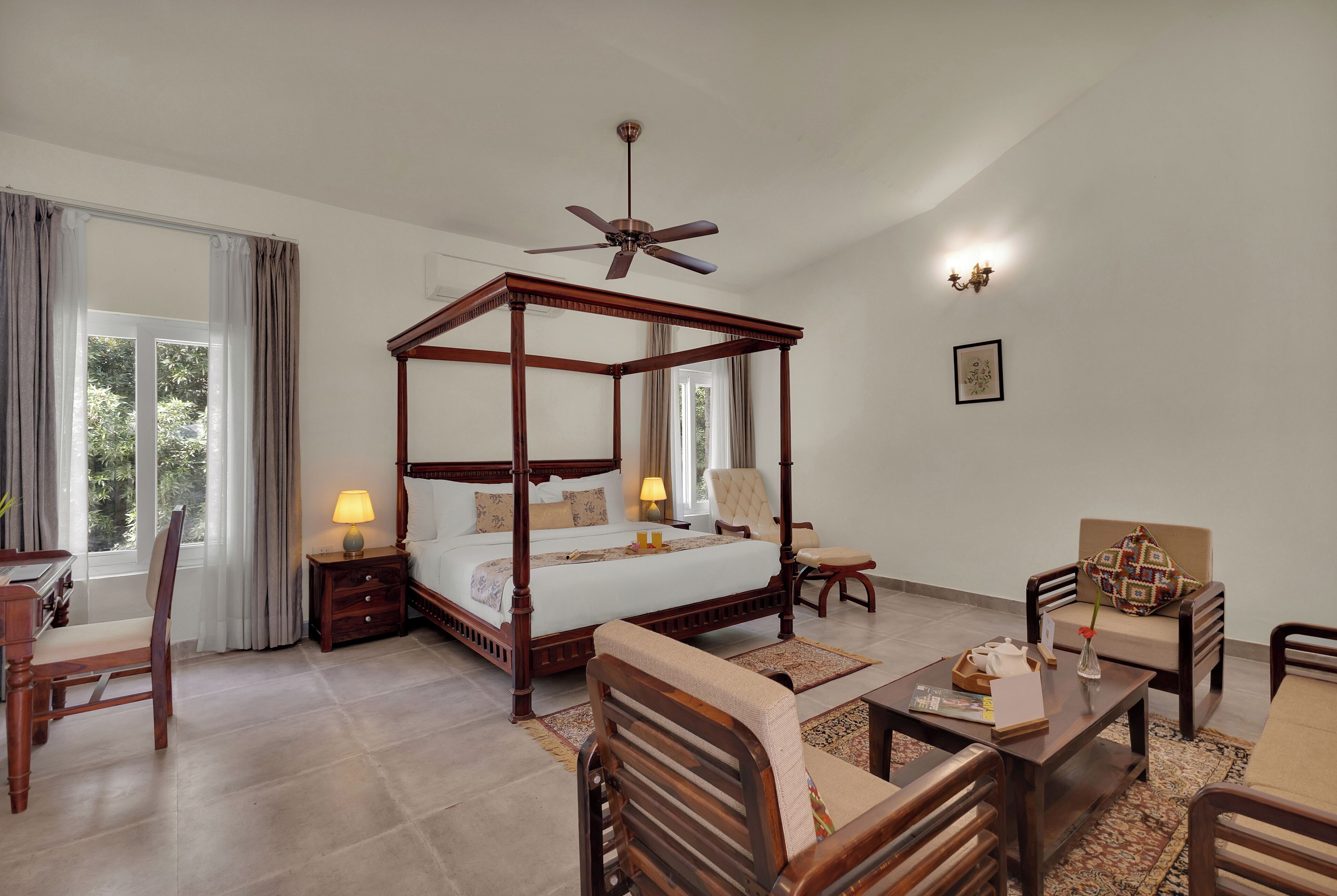 A four-poster bed with a ceiling fan, a desk with a chair, a sofa, and a small table with a vase.