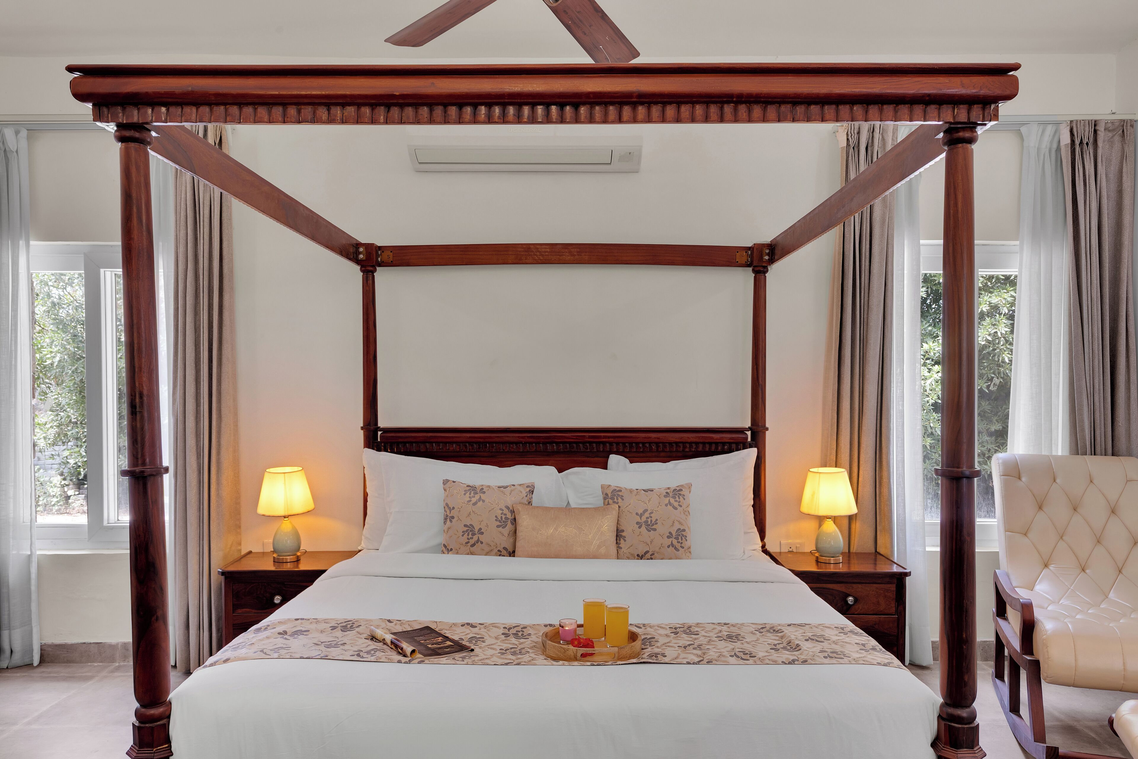 A four-poster bed with a wooden canopy, two bedside lamps, a tray with a book and a glass of juice, and a chair.